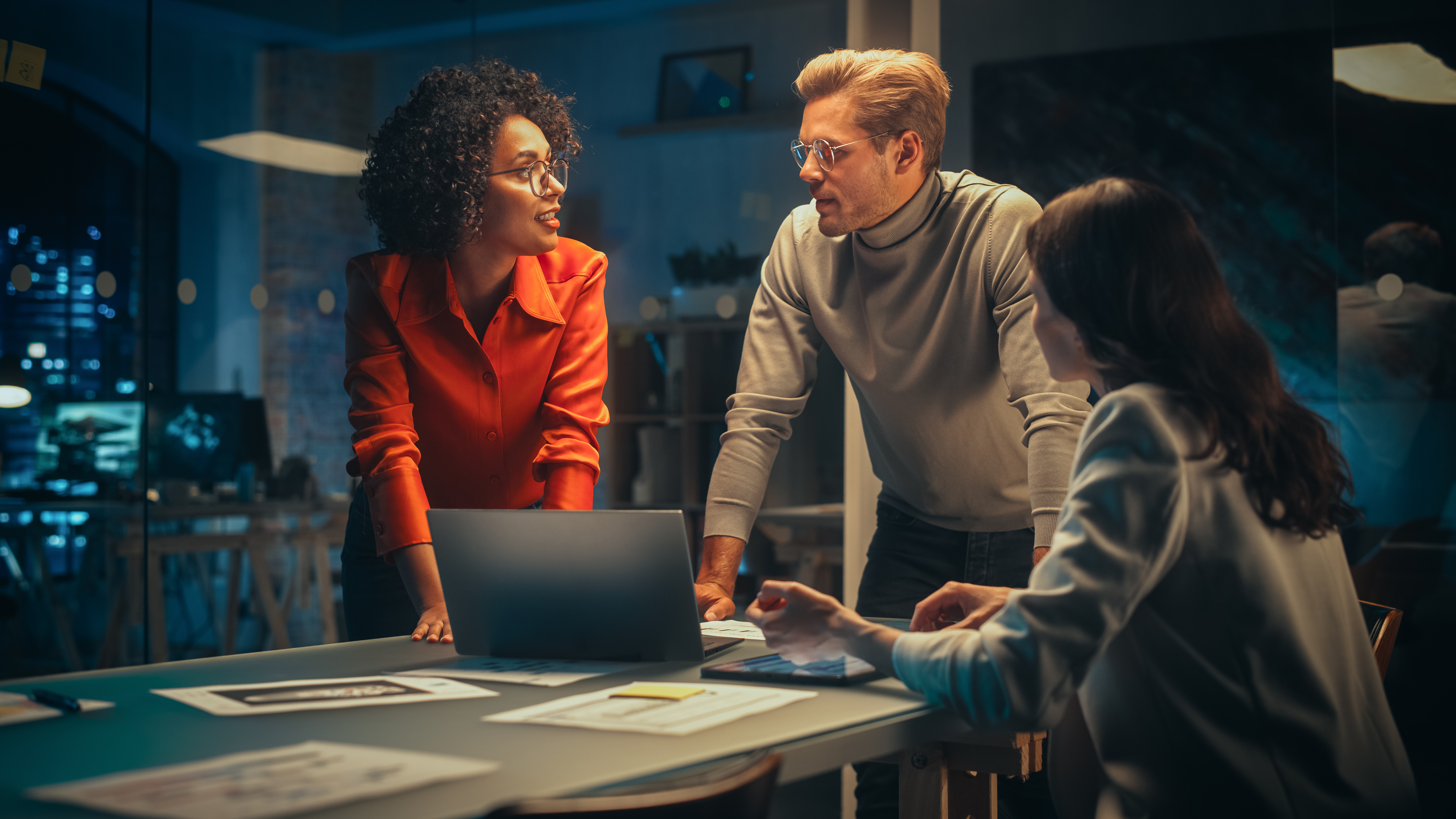 Small Creative Team Meeting In The Evening In Office Young Project Manager Bringing Her Research Paper, Focused Marketing Director Using Tablet Computer And Discussing The Analysis