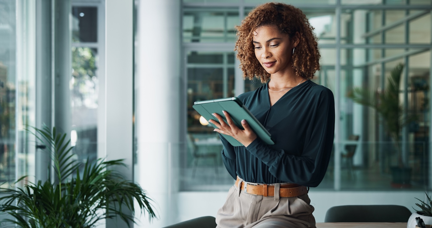 A woman uses a tablet while standing beside windows in a glass-walled office.