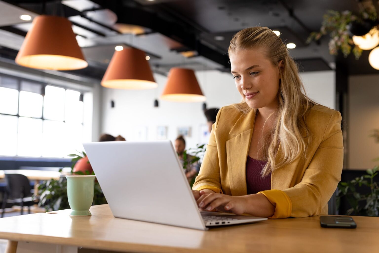 A woman in a mustard-colored jacket works on a laptop at a shared table.