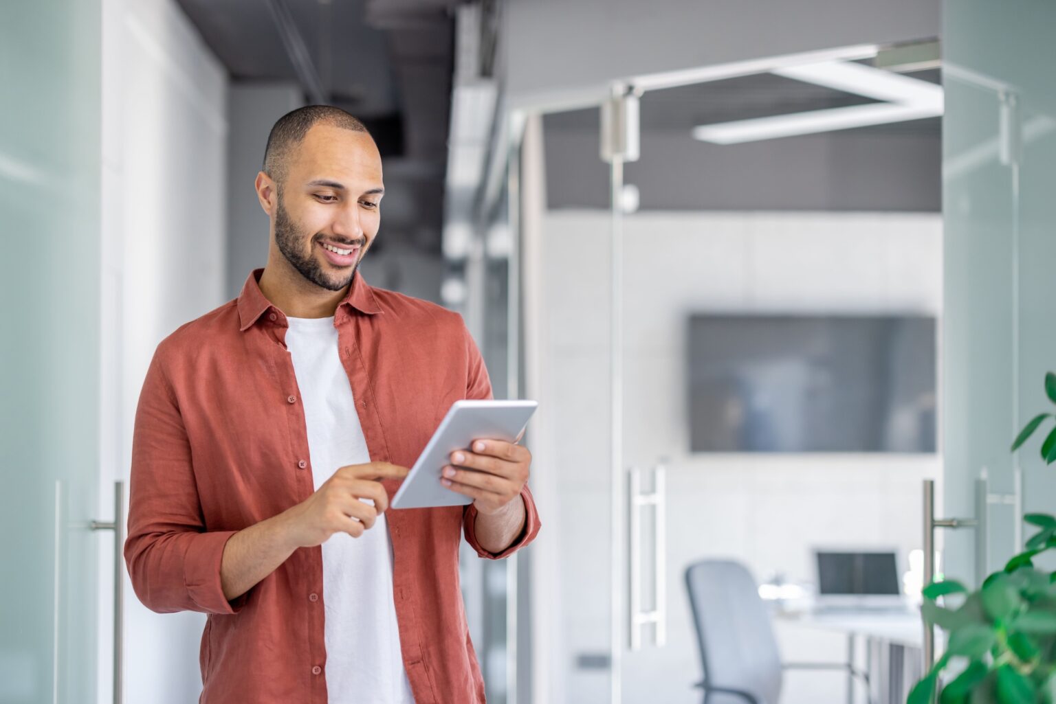 A smiling man in a red shirt uses a tablet while standing in an office.