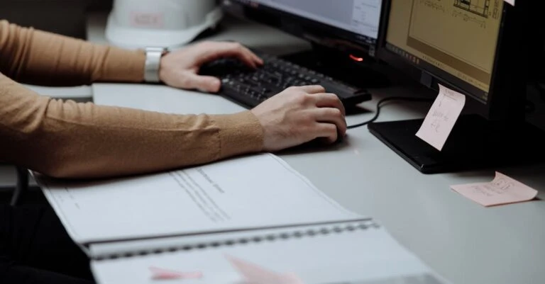 Hands using a keyboard and mouse at a dual-monitor workstation beside an open ring binder.