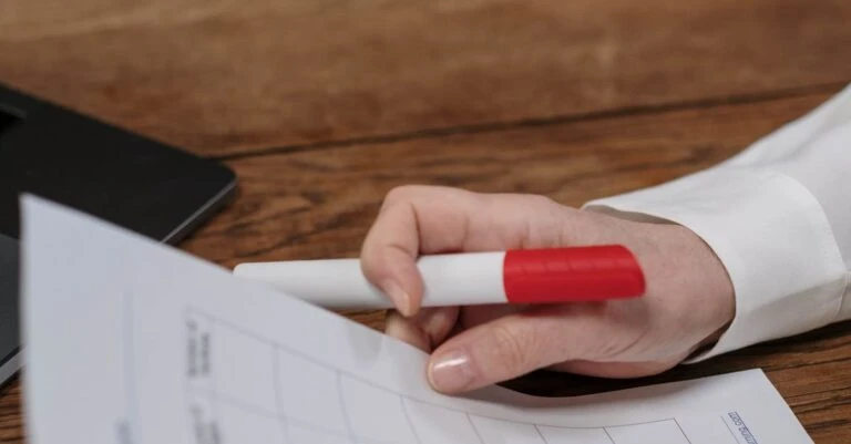 Hand holds a red-capped marker over printed paperwork on a wooden desk.