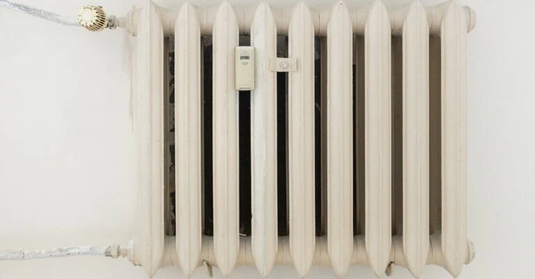 White ribbed radiator with exposed pipes mounted against a pale wall.