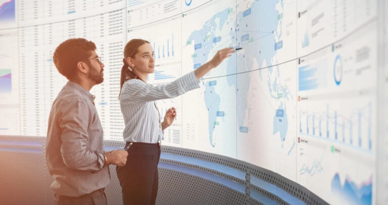 Indian Male Data Scientist And Caucasian Female Business Manager Talking In Front Of Big Digital Screen With Graphs, Charts And Map In Monitoring Office Diverse Colleagues Discussing Objectives
