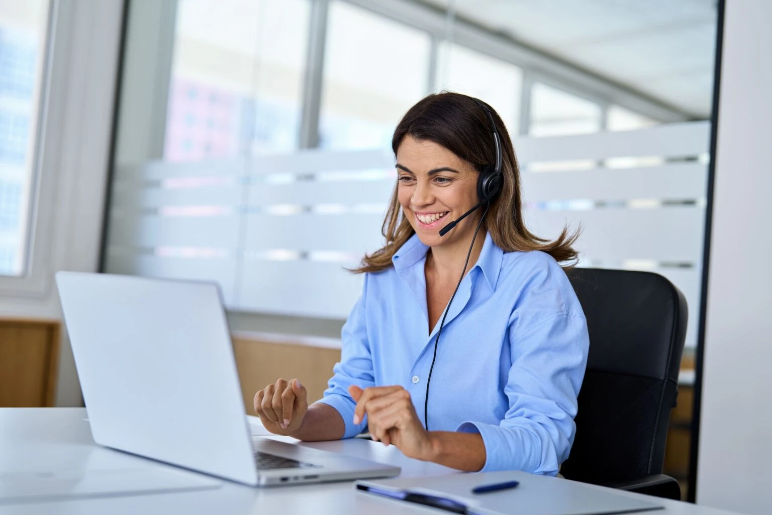 A woman wearing a headset smiles while typing on a laptop at an office desk.