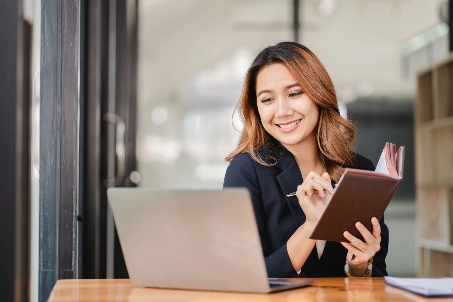 A smiling woman holds an open notebook while seated beside a laptop.