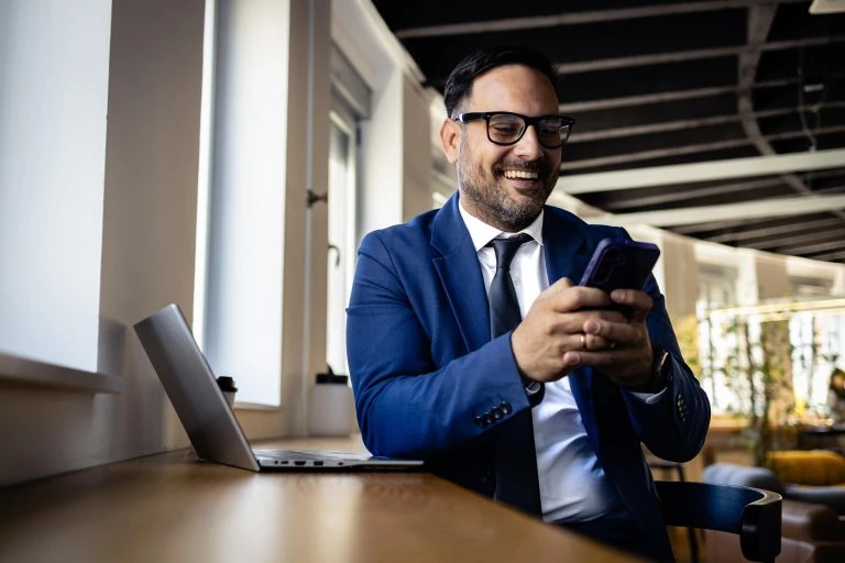 Businessman smiling, texting on smartphone, using laptop in modern office, finding business success.